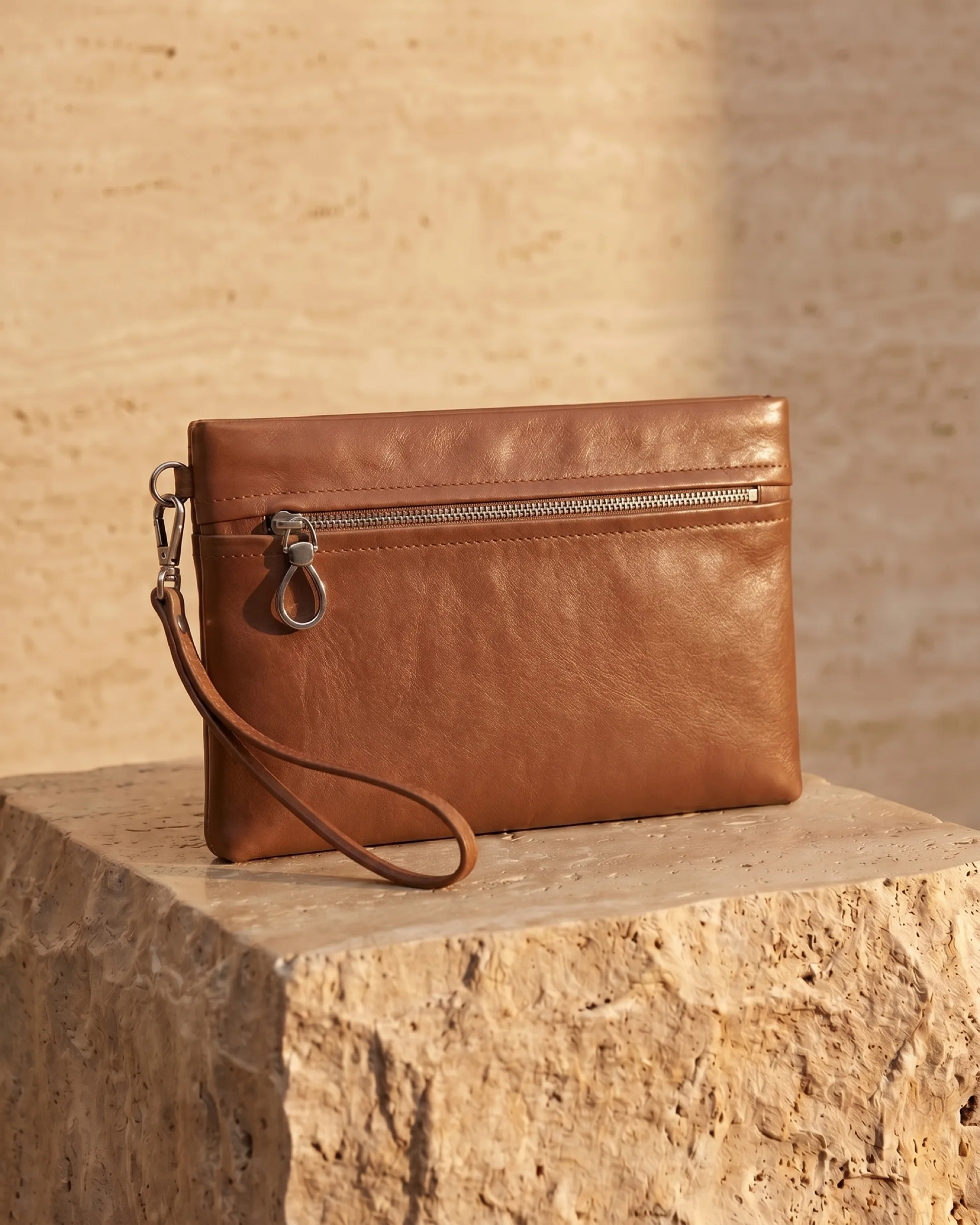 Hugo Caesar Tassi Viminia small leather evening clutch in Cuoio coffee-brown, top-zip with silver pull and wrist strap, on a warm travertine pedestal