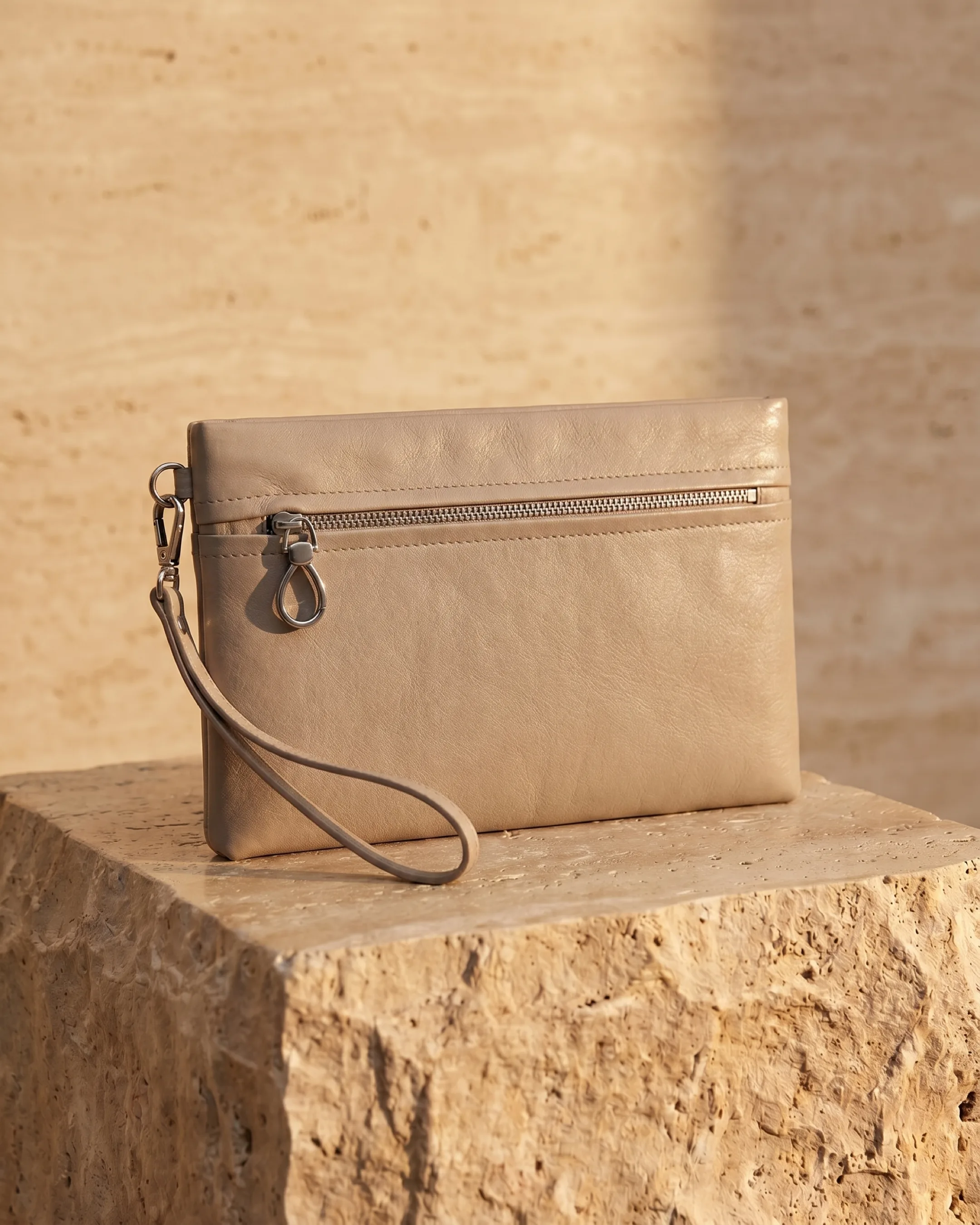 Hugo Caesar Tassi Viminia small leather evening clutch in Travertine pale stone, top-zip with silver pull and wrist strap, on a warm travertine pedestal