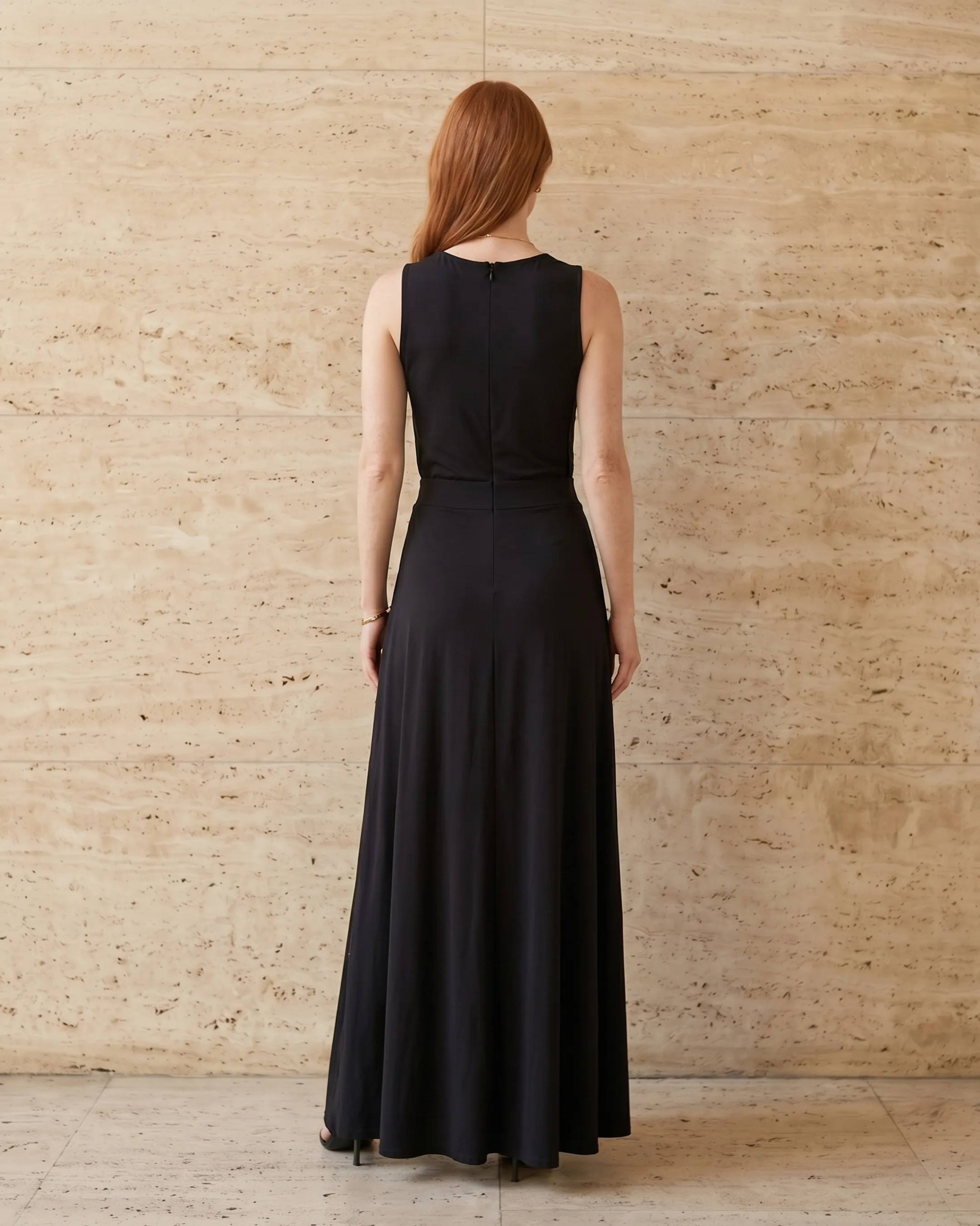 Full-length rear view of the Hugo Caesar Tassi Vipsania draped modal column gown in Basalt black, seen from behind, showing the back of the dress, the sleeveless high neck, high gathered waist and fluid modal-jersey column falling to a soft train in a Roman travertine studio